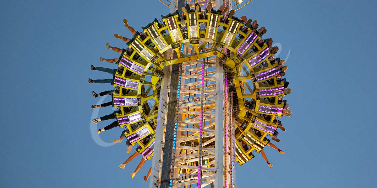 Large drop tower ride with illuminated seats and passengers’ legs extended, showing the excitement and safety features of a custom drop tower in operation at night.