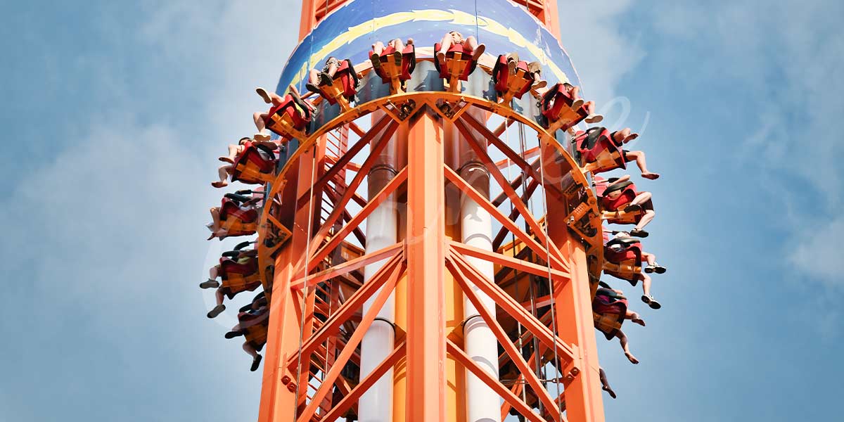 Close-up of orange drop tower ride with riders at the top, highlighting the height, secure seating, and engineering of a modern thrill ride.