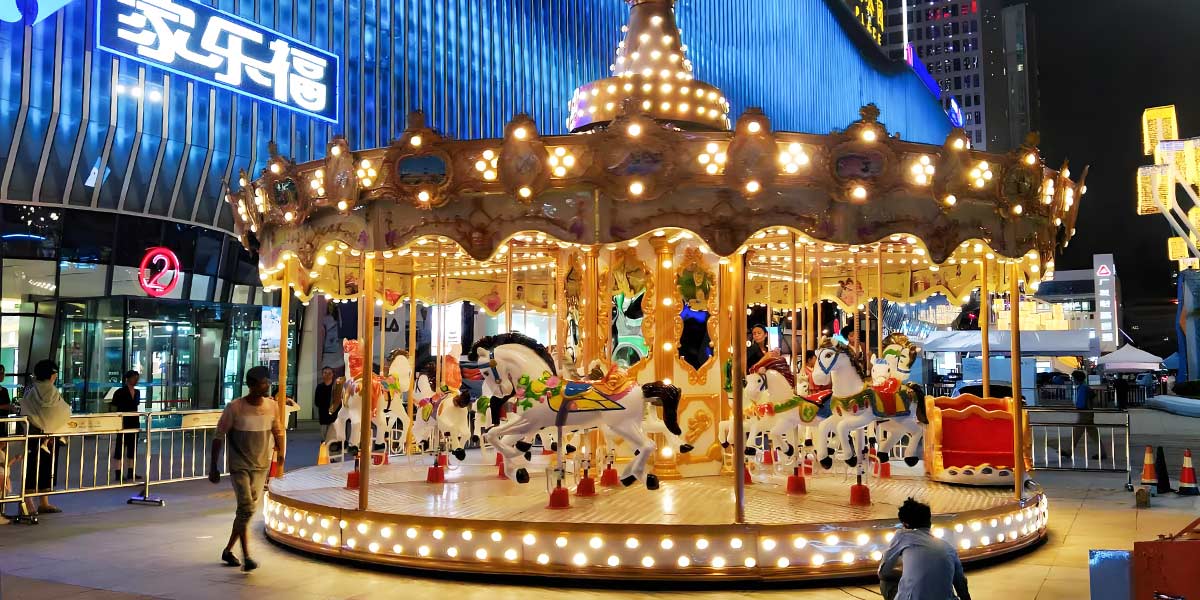 Paseo en carrusel luminoso con caballos blancos por la noche en un concurrido centro comercial, que atrae visitantes y añade un ambiente vibrante a la zona.