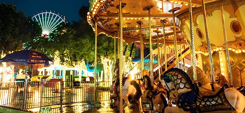 A 2 story carousel in an amusement park at night, illuminated by bright lights, with other attractions like a ferris wheel in the background