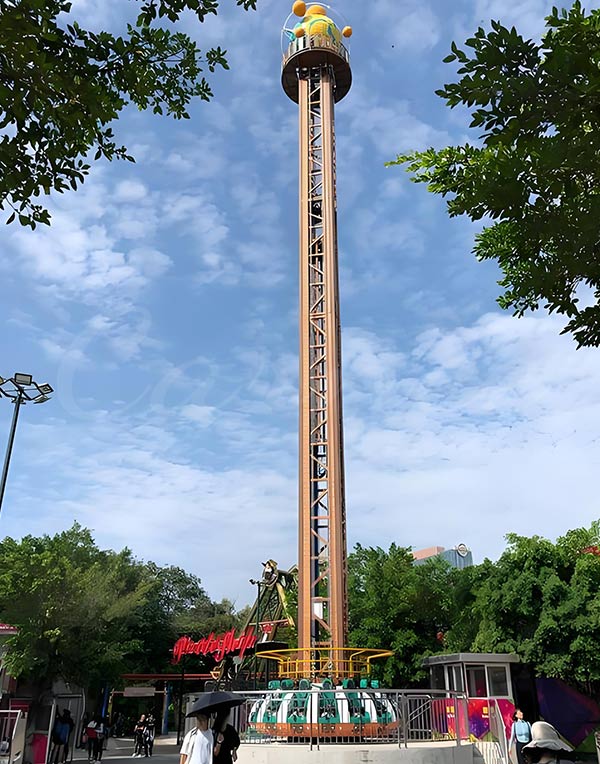 Drop tower amusement ride with a towering structure at an amusement park, surrounded by trees and greenery.