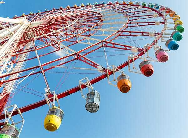 Uma grande roda-gigante com gôndolas coloridas em um céu azul. Conhecida como Big Wheel ou Observation Wheel no Reino Unido, ela serve como um passeio âncora altamente lucrativo para parques de diversões.