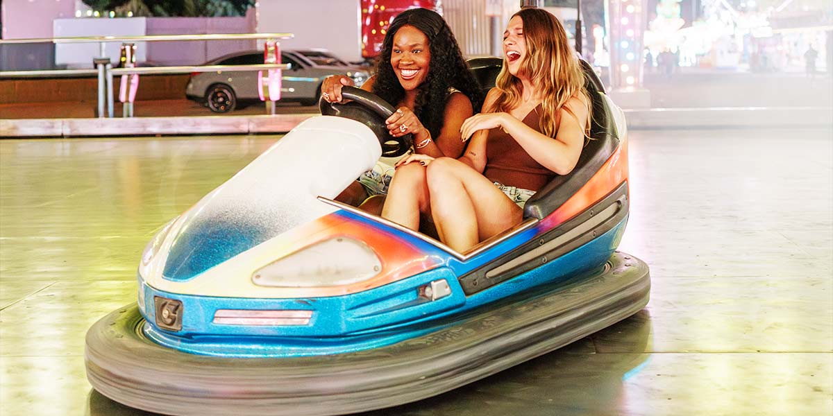 Duas mulheres jovens rindo enquanto andam de Dodgems, comumente conhecidos como Bumper Cars nos EUA, destacando o potencial de alto lucro dos parques de diversões.