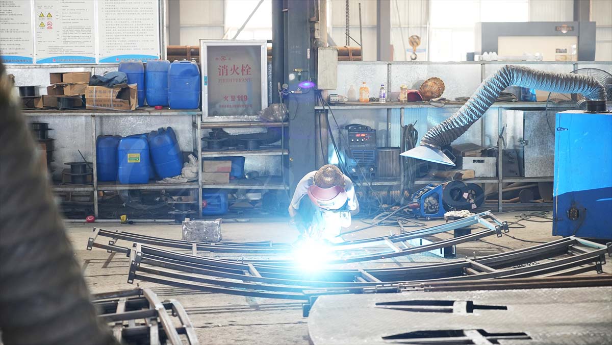 Un ouvrier qualifié portant un masque de protection soude une section de rail métallique incurvée dans une usine de fabrication de manèges en Chine. La lumière bleue vibrante de l'arc de soudage est proéminente. Dans l'arrière-plan détaillé de l'atelier, on peut voir des étagères industrielles, des tonneaux bleus, un panneau de signalisation (bouche d'incendie) et des matériaux métalliques, qui témoignent de la qualité de la fabrication des attractions des parcs d'attractions.